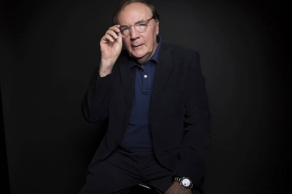 Author photo of James Patterson, he's holding his glasses on his face with his right hand, sitting on a stool against a black background