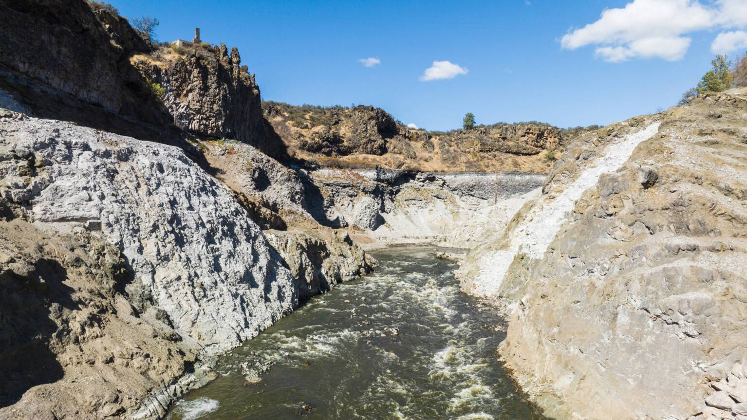 A river flows through a rocky gorge with steep limestone and basalt cliffs on both sides, scattered vegetation on the hillsides above, and clear blue sky with white clouds.