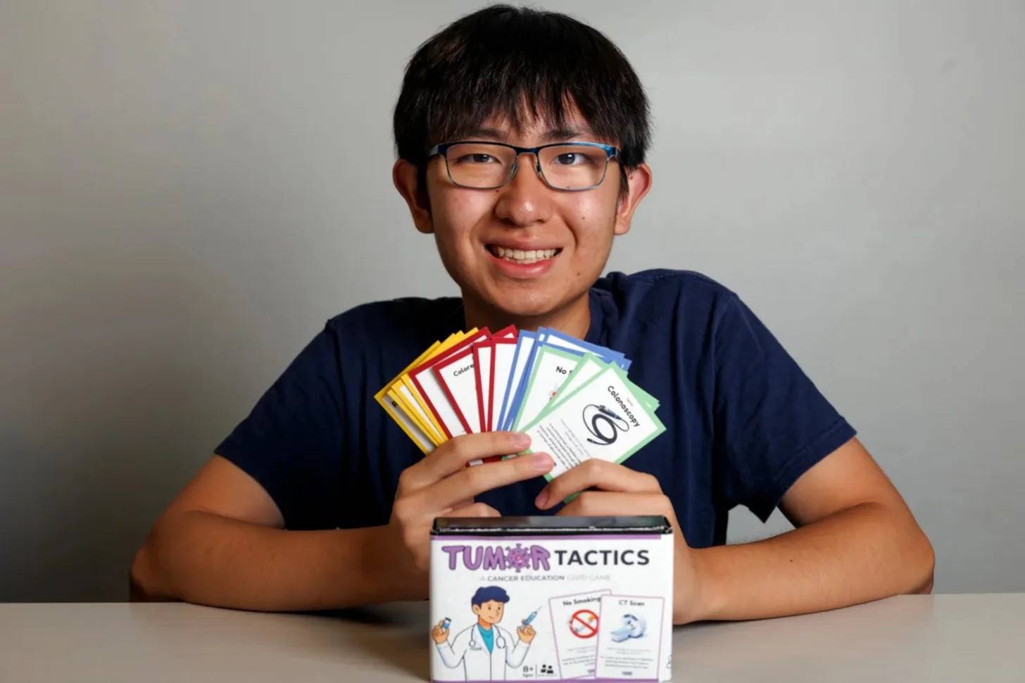 A young person wearing glasses and a dark blue shirt holds fanned-out game cards above a board game box labeled "Tumor Tactics" with cancer education imagery.