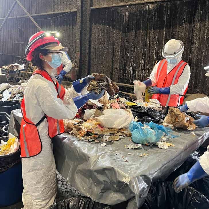 Two people wearing head lamps, masks, orange vests, and white jumpsuits looking through trash