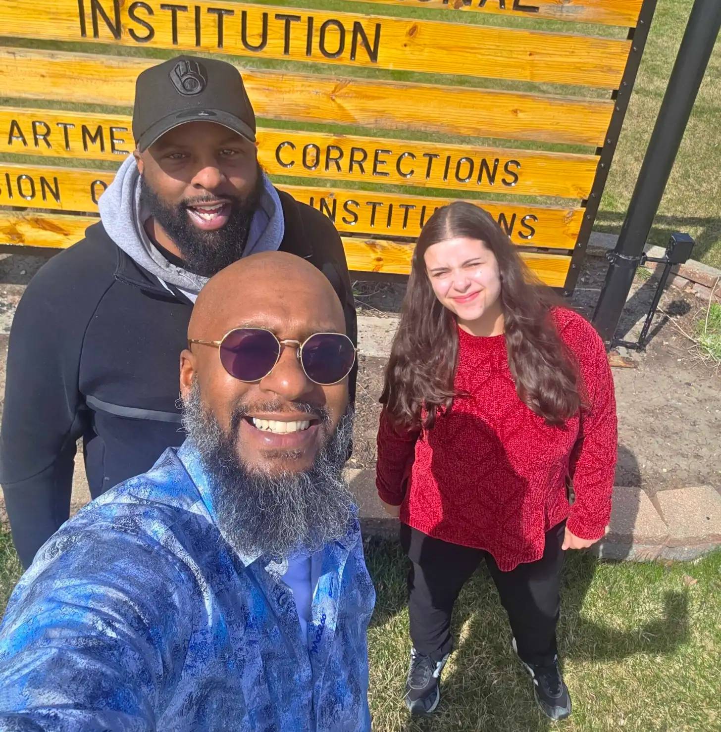 Three people stand together outdoors in front of a yellow wooden structure with partial text reading "CORRECTIONS" and "INSTITUTION" visible. The man in front wears a blue velvet jacket and round purple-tinted sunglasses. The man behind wears a black cap and dark hoodie. The woman on the right wears a red knit sweater. All three are smiling at the camera.