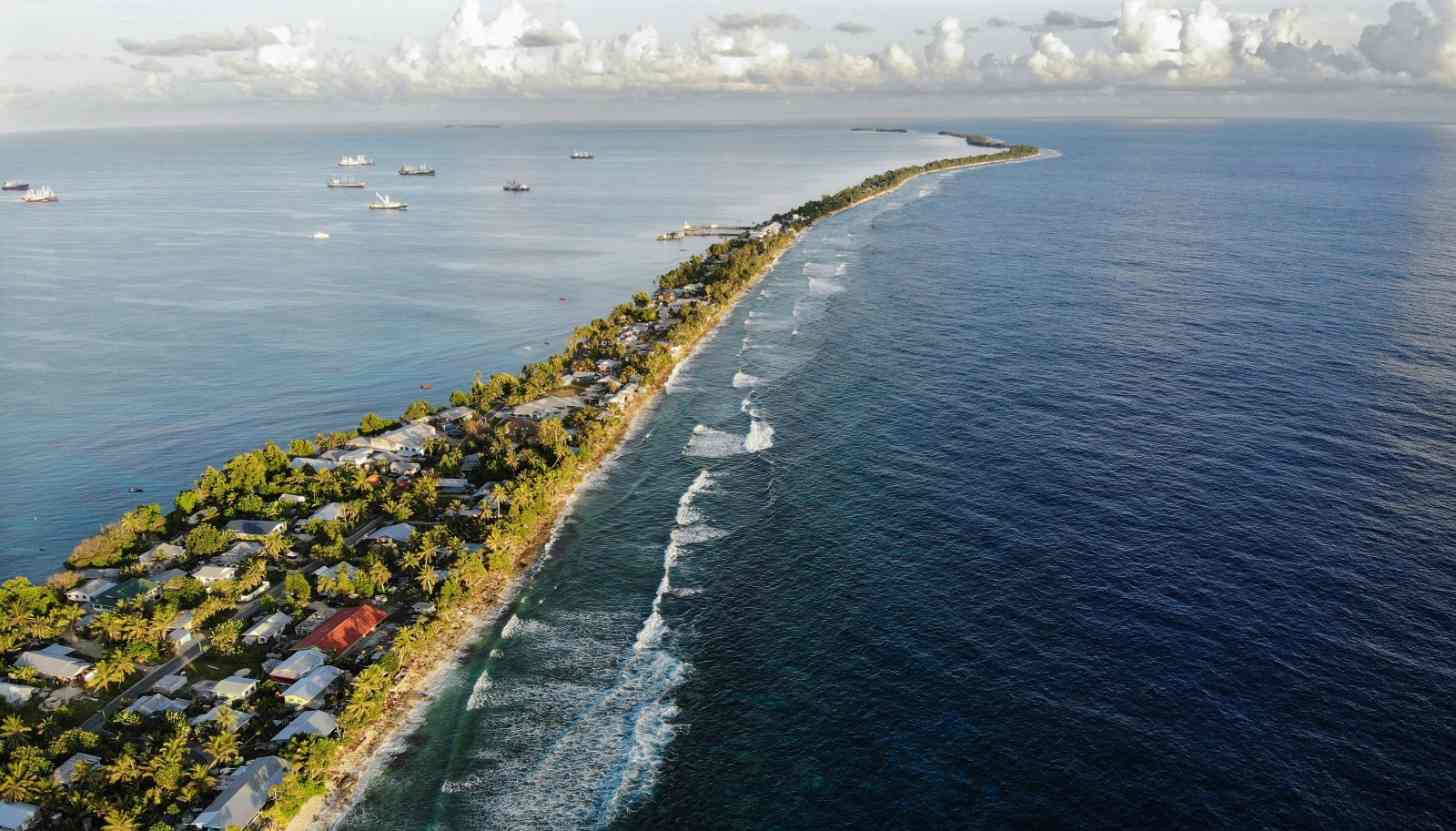 Aerial view of a narrow island with palm trees and buildings along its shore, separating a calm lagoon on the left with anchored boats from rougher ocean waters on the right with visible white-capped waves.