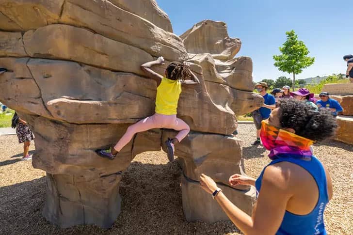 An adult wearing a blue tank top and colorful face covering spots a young girl climbing on a small rock formation at a park