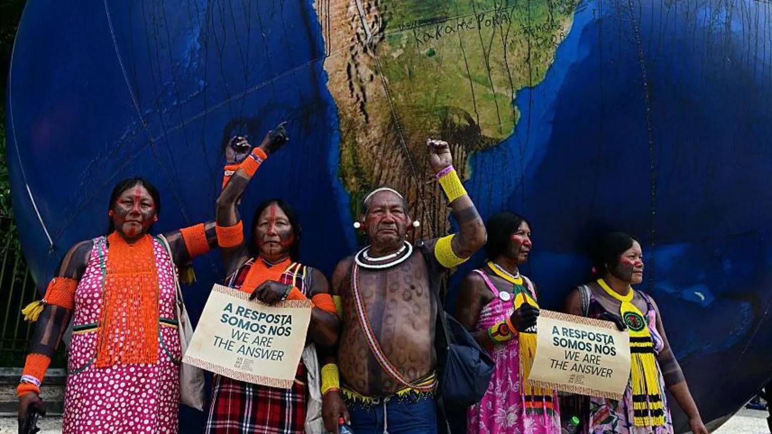 Five Indigenous protesters in front of a massive globe holding signs that read "we are the answer"