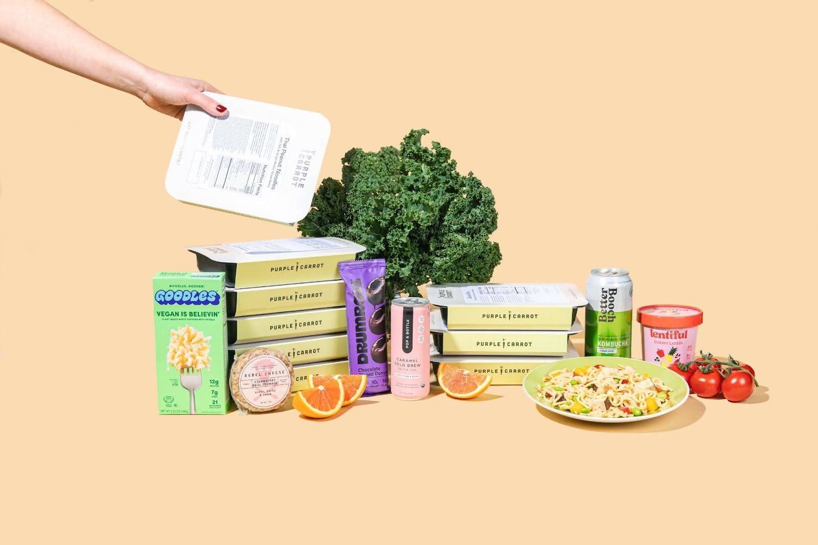 A box of goodles, pre-packed Purple Carrot meals, a can of Better Booch, a plate of food, and more artfully displayed on a light orange background with a hand reaching from off-screen to grab a packaged meal