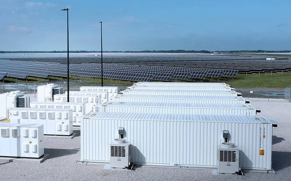 White containerized battery storage units and electrical equipment sit on pavement in front of a large solar panel farm under a blue sky.