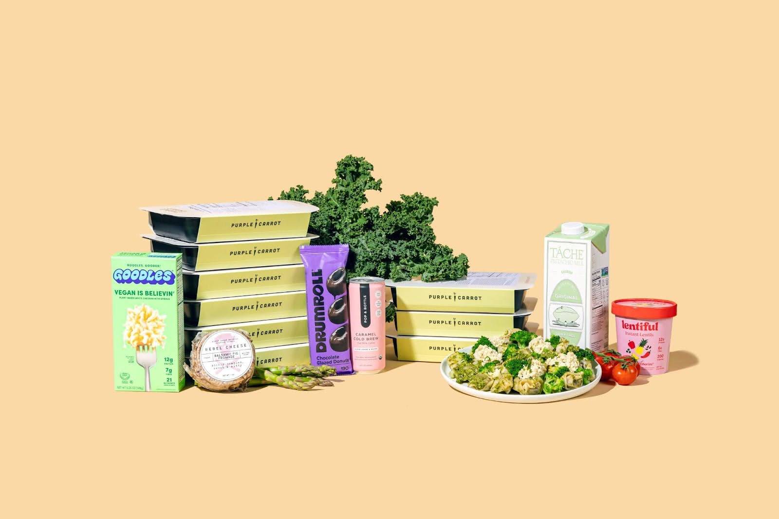 Pre-made Purple Carrot meal boxes stacked next to a box of mac-n-cheese, a package of cheese, kale, a plate of food, and other grocery items on a light orange background