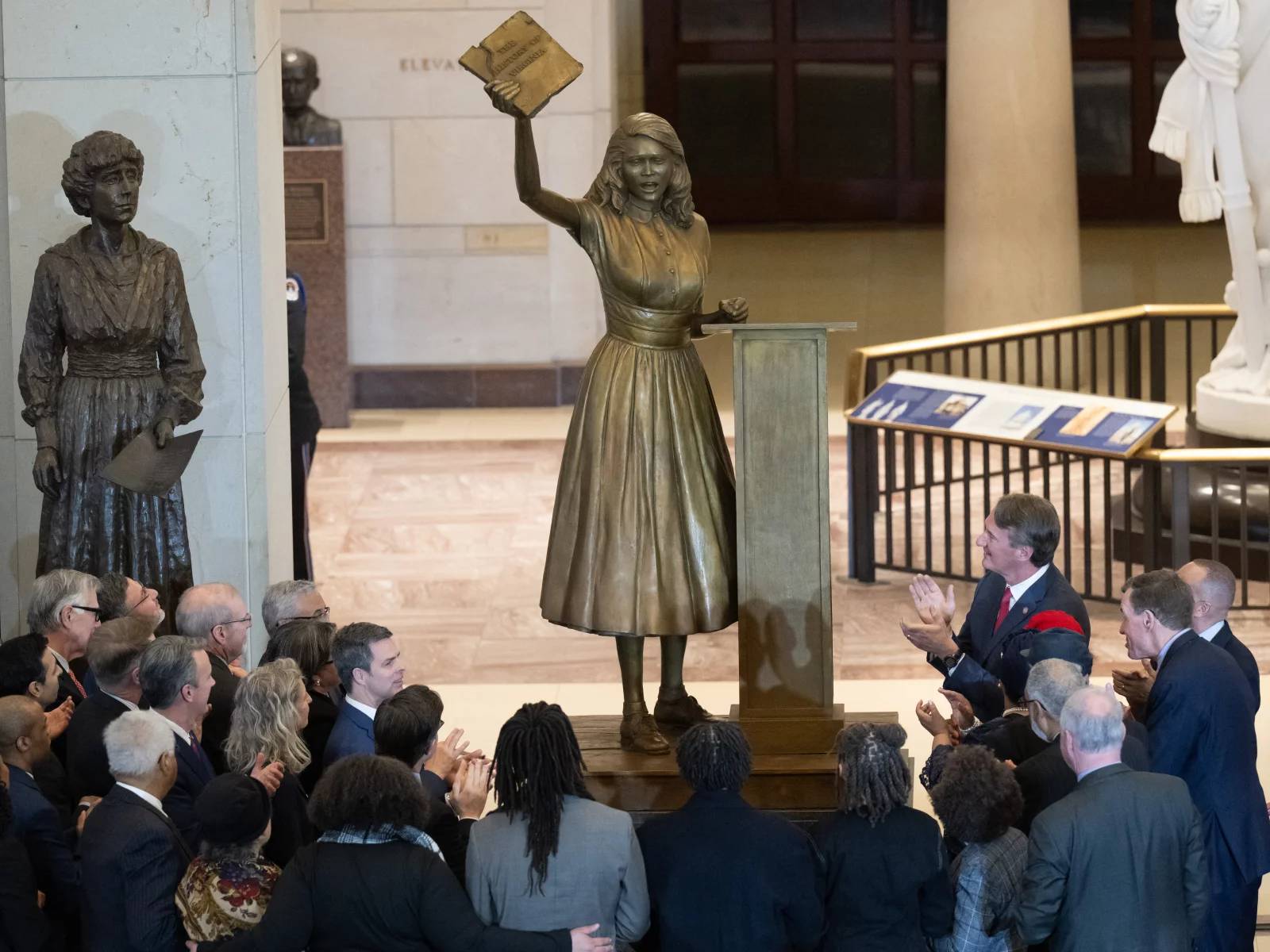 Bronze-colored statue in Capitol with people gathered around it clapping