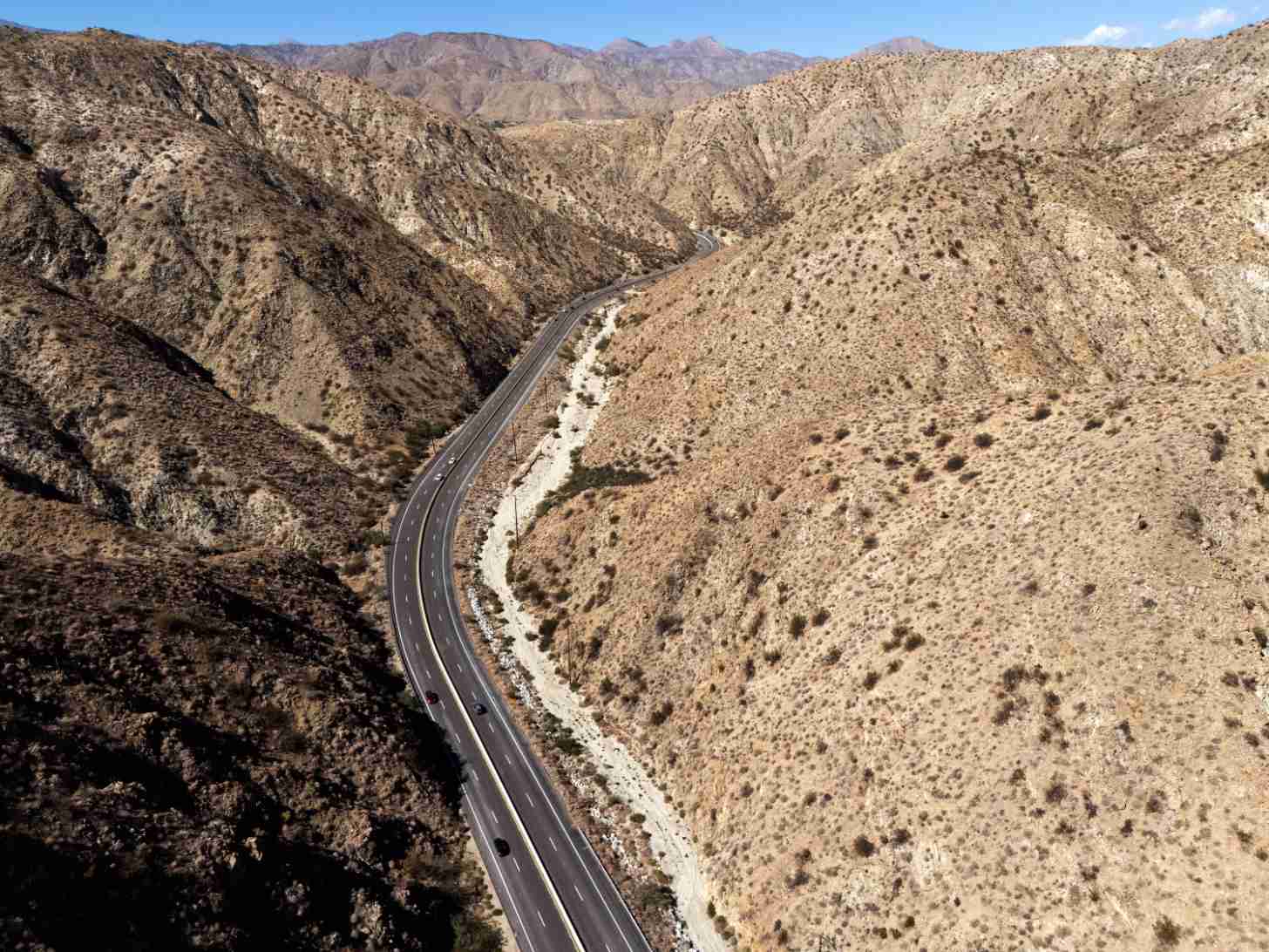 Aerial view of a paved highway winding through a desert canyon with rocky mountainsides and a shallow stream running alongside the road.