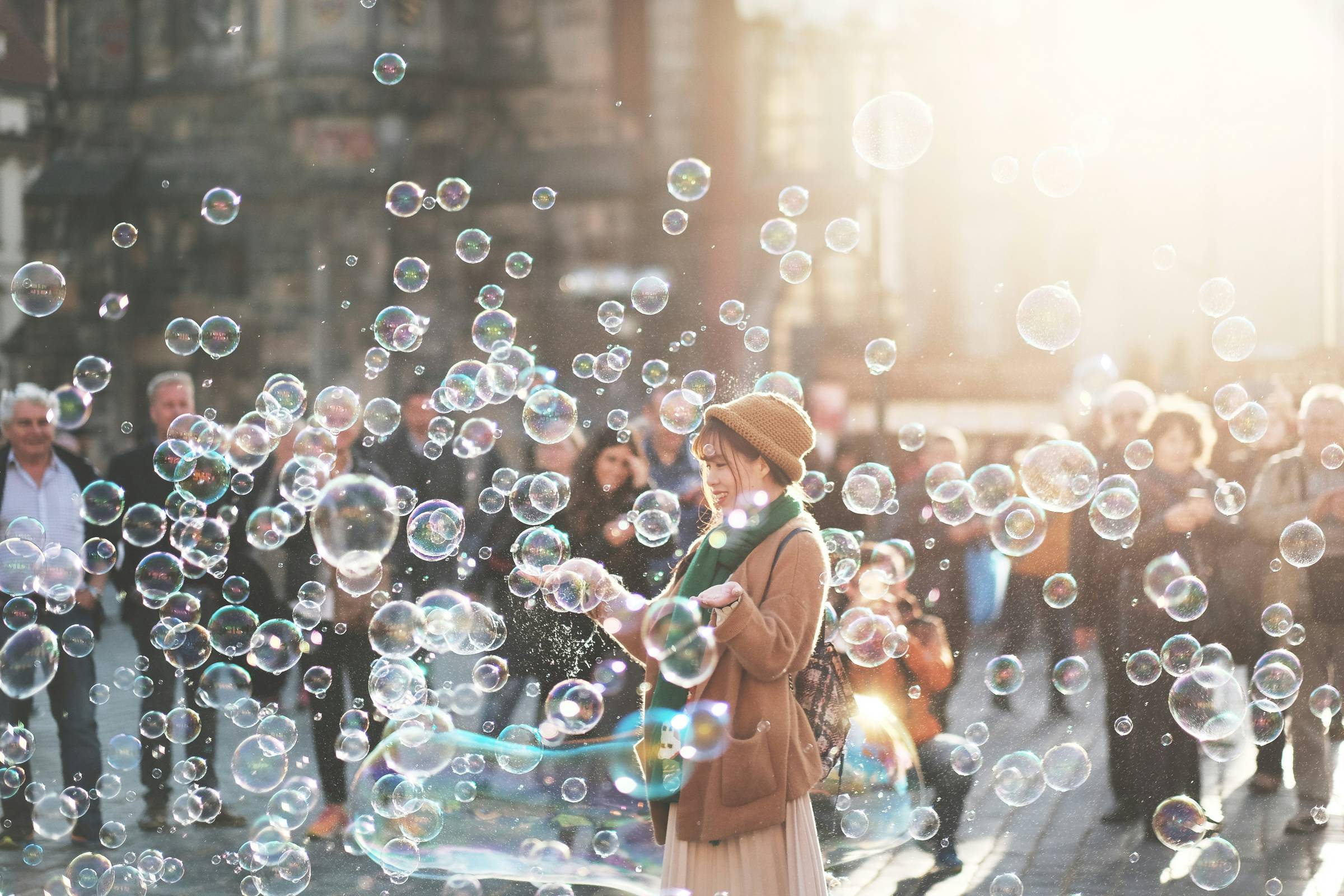 A person in a tan coat and brown hat blows bubbles in a crowded urban street, with numerous translucent soap bubbles floating through the air around them and spectators visible in the background.