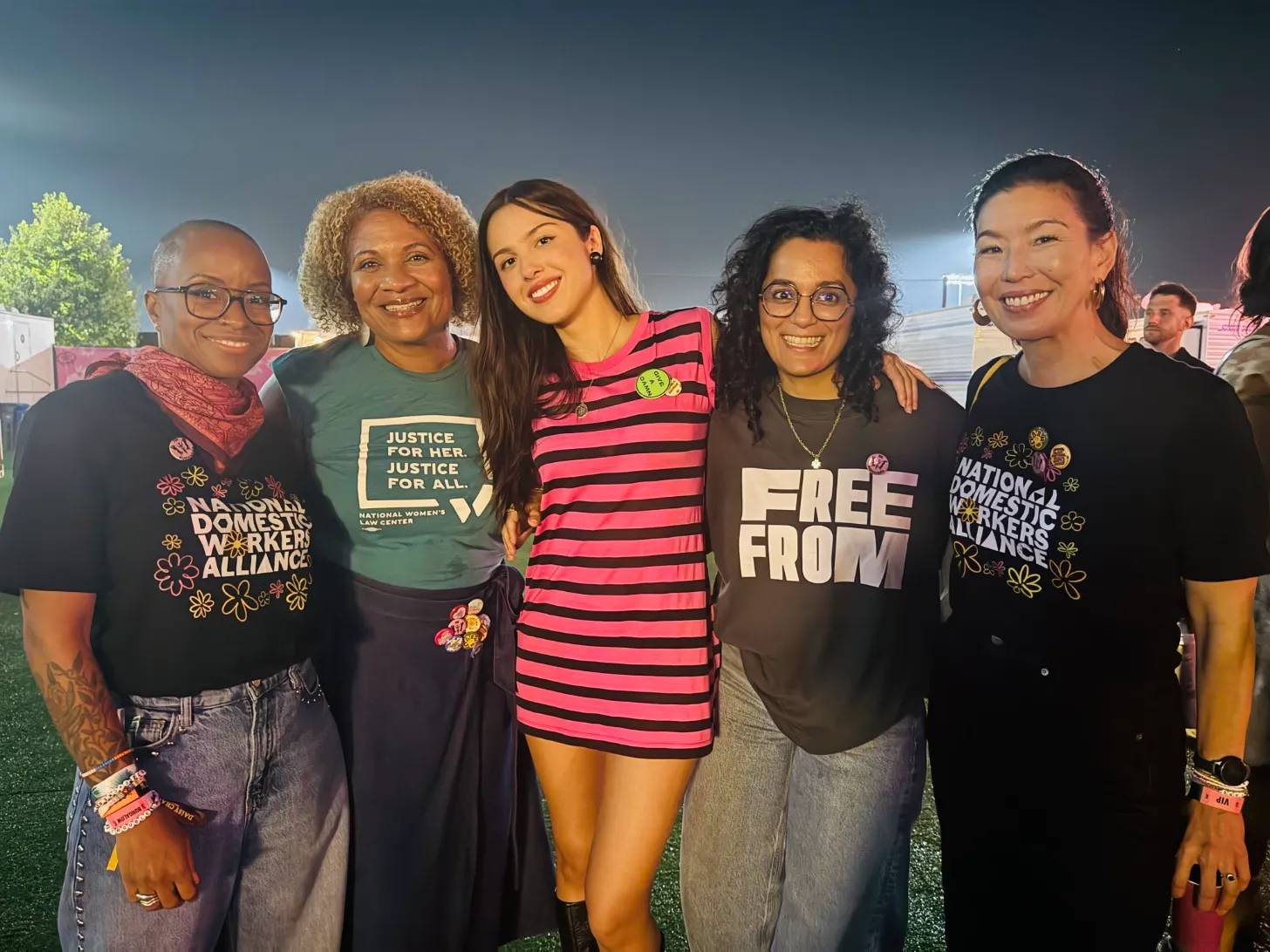 Five people stand together outdoors at dusk, smiling at the camera. Their shirts display text including "National Domestic Workers Alliance," "Justice for Her. Justice for All," "Free From," and "National Domestic Workers Alliance."