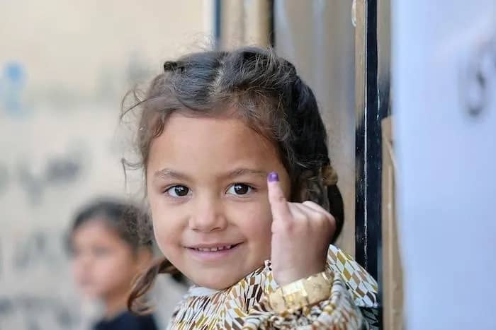 A small child with braids in her hair looking at the camera and holding up her pinkie, which has a purple marking on the end of it