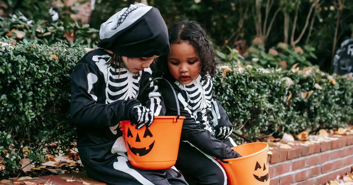 Two children wearing skeleton costumes sit on a brick pathway surrounded by hedges, holding orange jack-o'-lantern buckets.