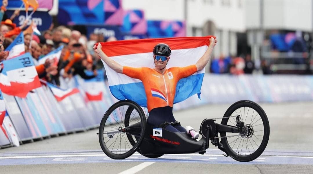 An adaptive biker holds a flag for France behind them with people cheering on the sidelines