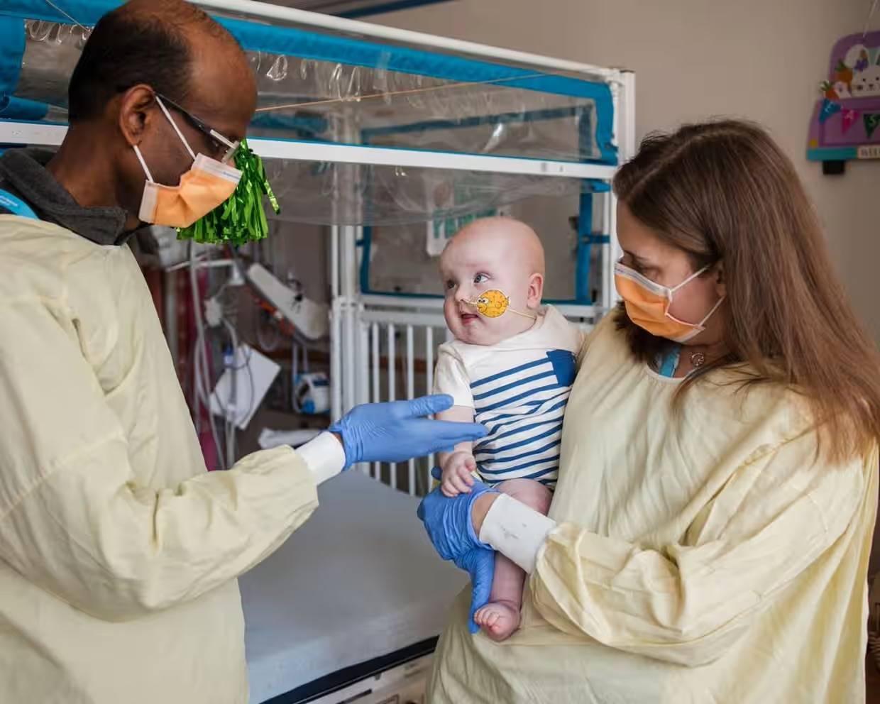 A woman wearing yellow medical dressing holding a baby looking at a man wearing yellow medical dressing