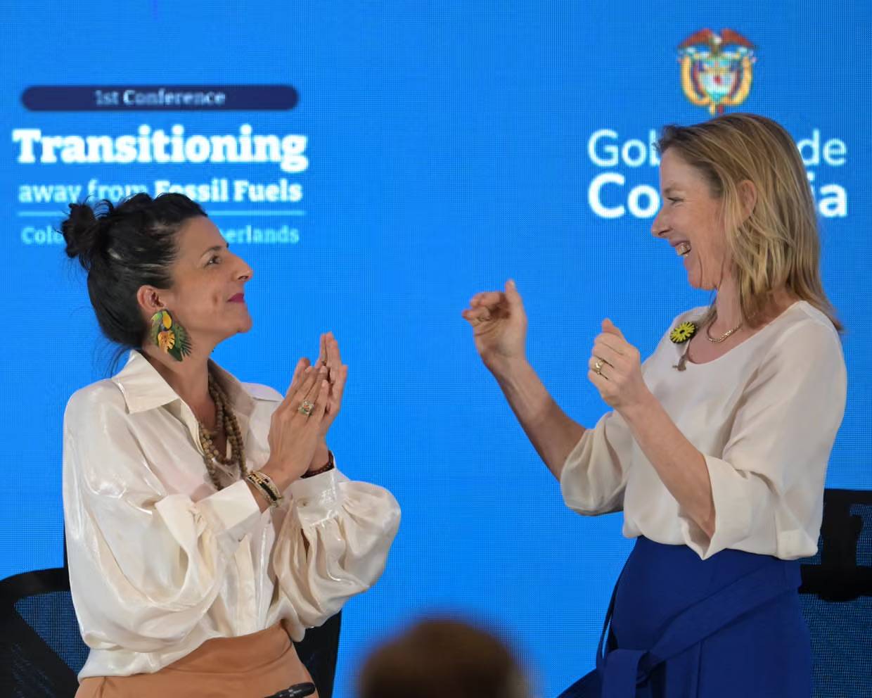 Two women smiling, clapping, and looking at each other in front of a blue bright background with text on it