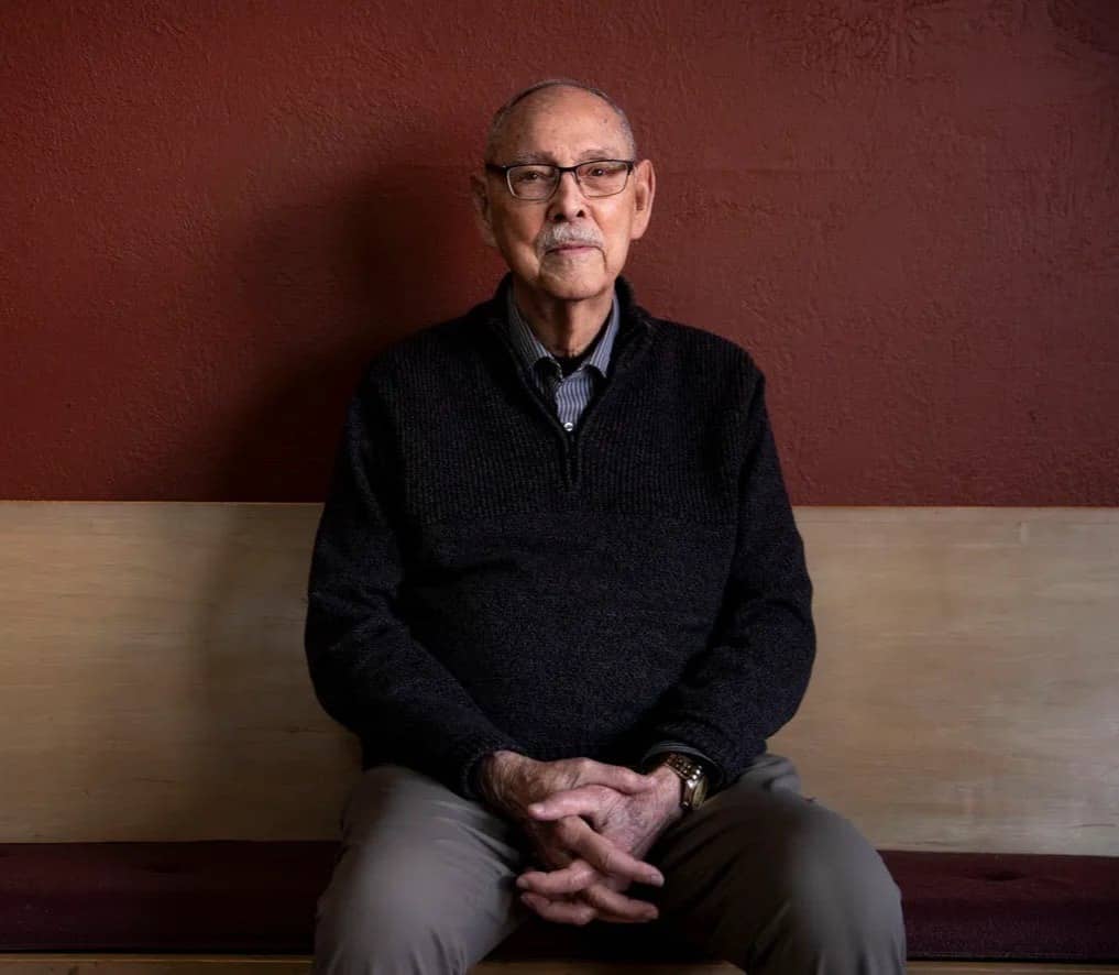 A profile image of Ron Post sitting on a bench against a wooden and dark red-painted background