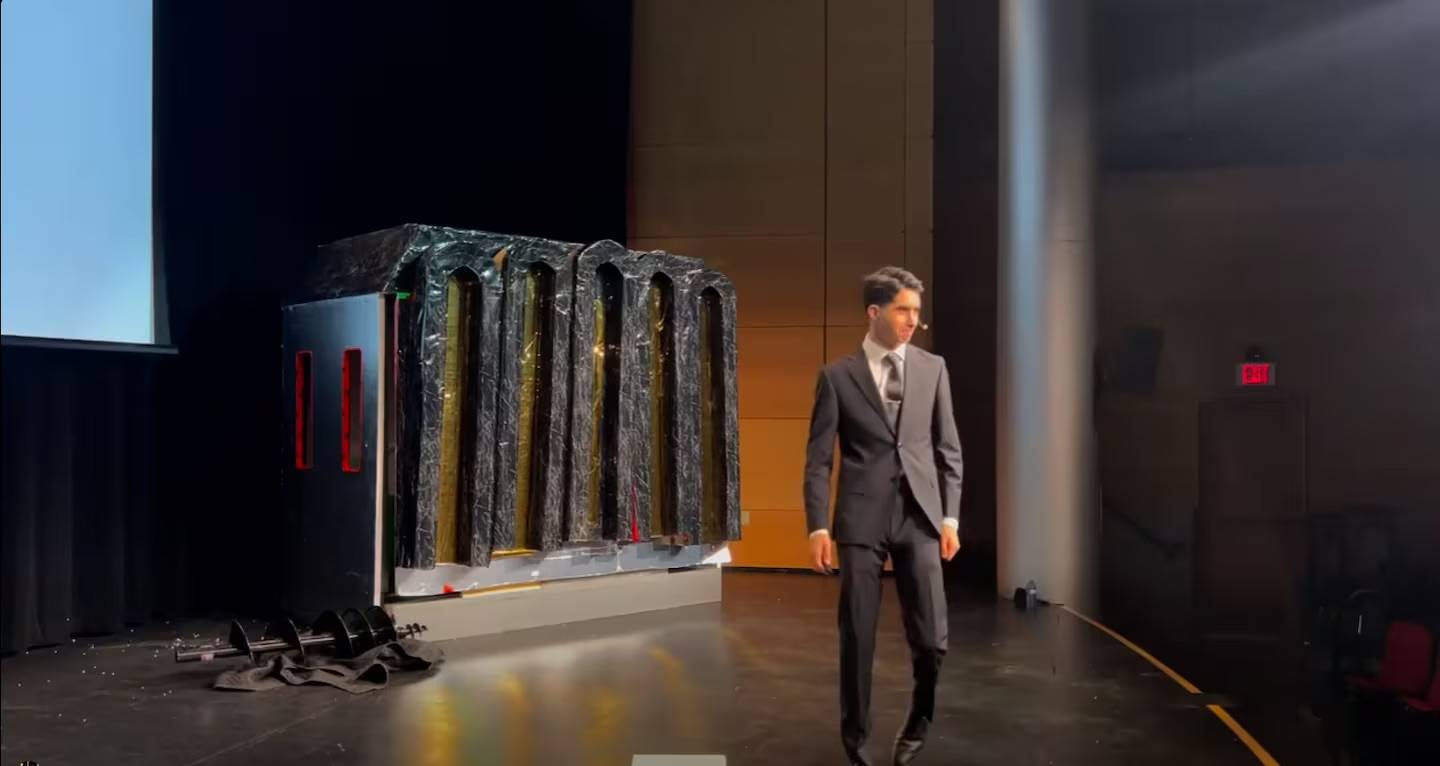 A person in a suit and tie on stage with a model structure behind them