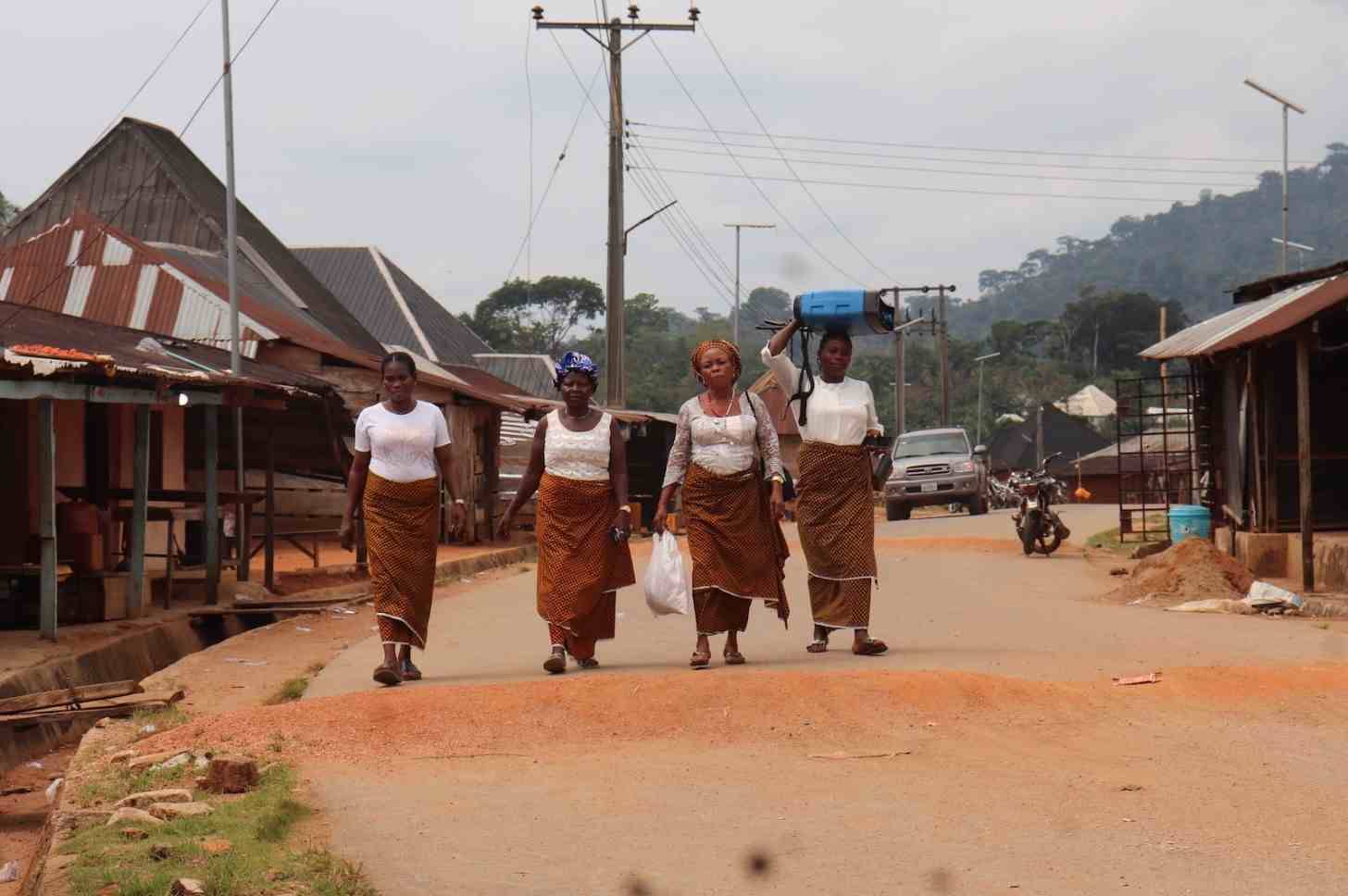 Four women wearing white tops and brown-orange skirts walking down a dirt road with structures on either side