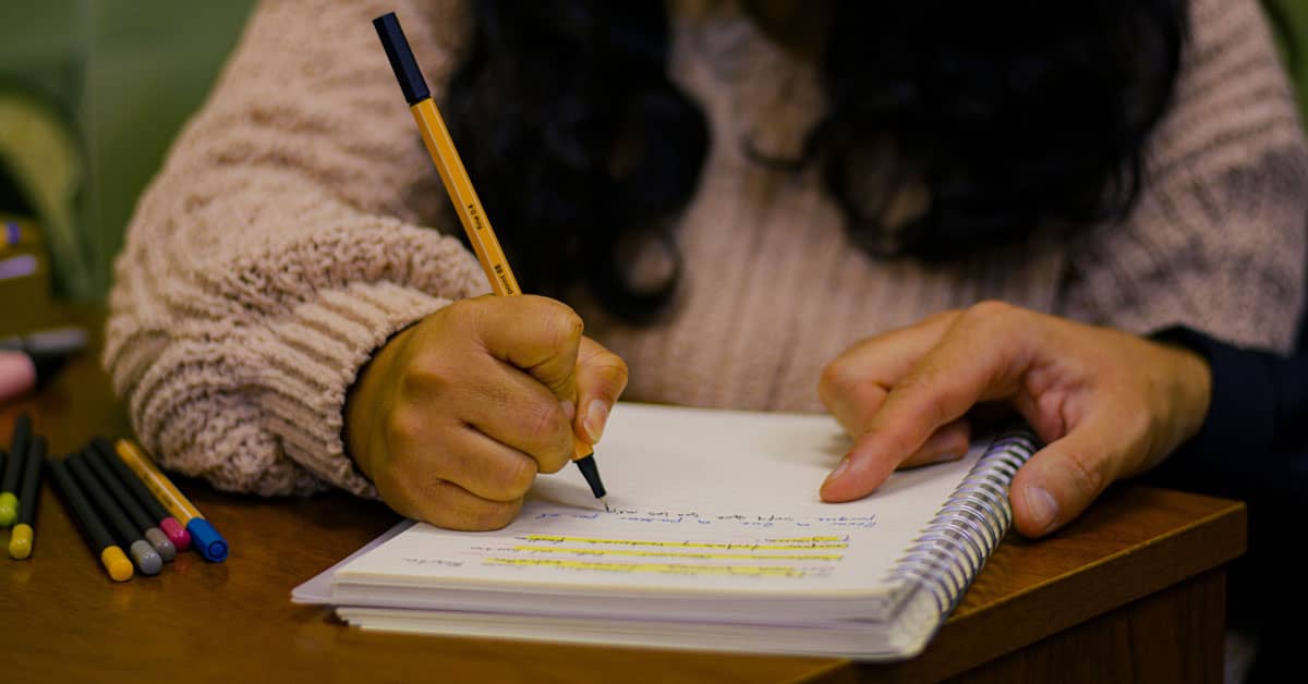 A child's hand holds a pencil while writing in a spiral notebook on a wooden table, with colored markers visible to the left.