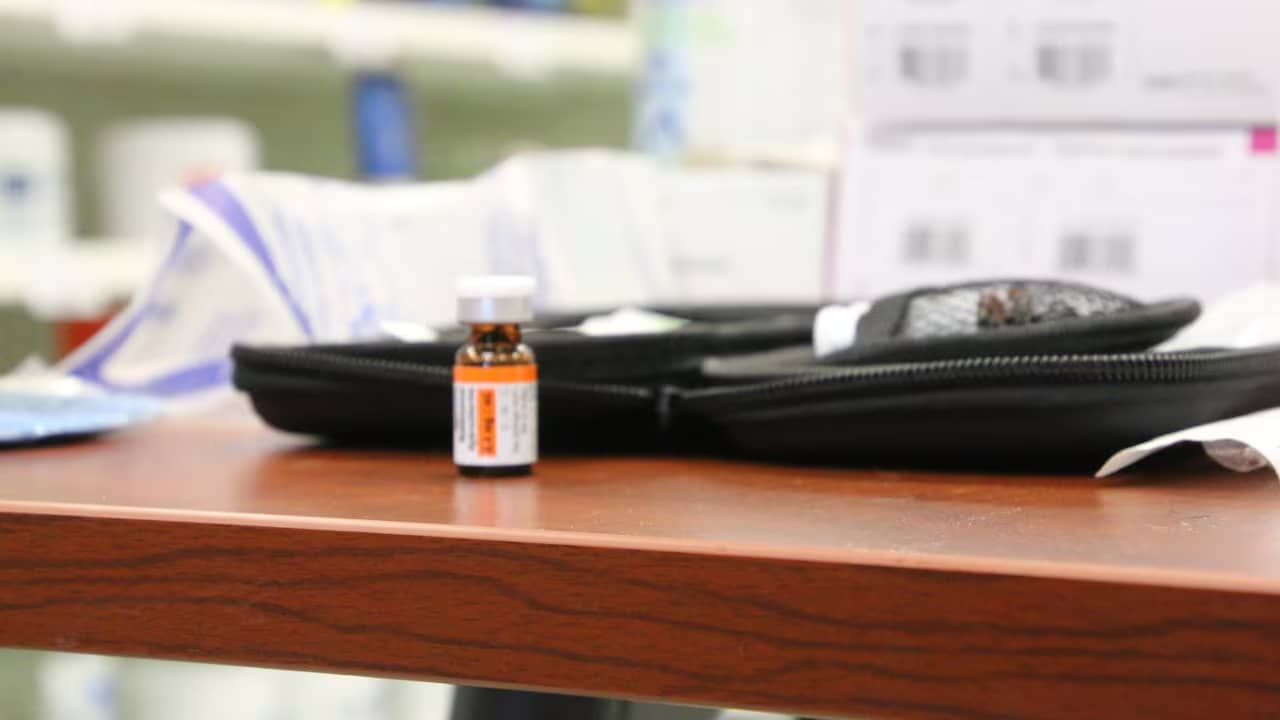 A small amber bottle with an orange and white label sits on a wooden desk among office supplies including a black computer mouse and papers, with blurred office furniture in the background.