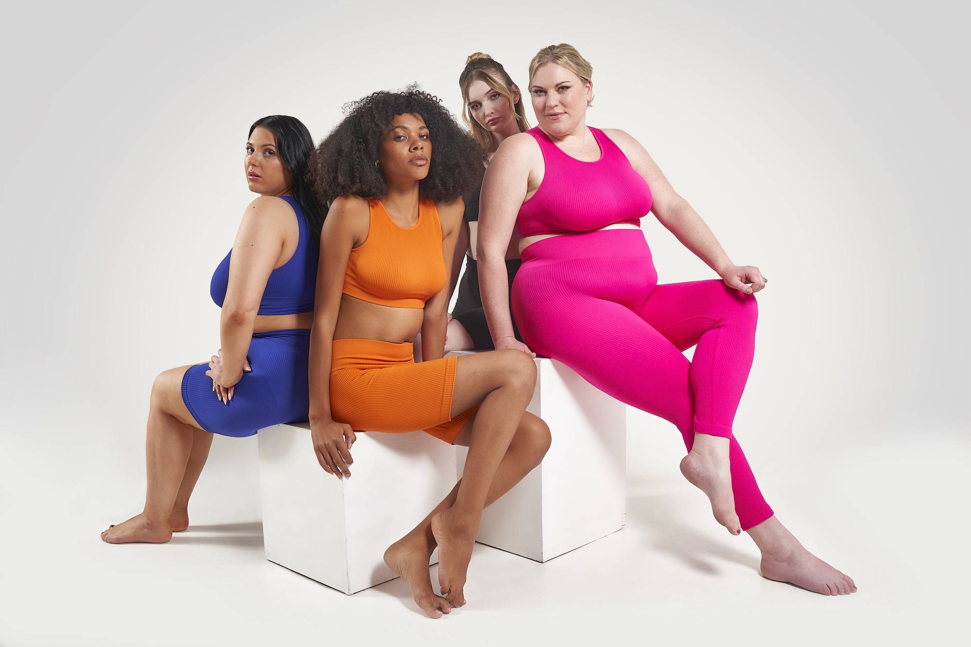 Four people sitting on two white blocks wearing blue, orange, black, and magenta athleisure sets