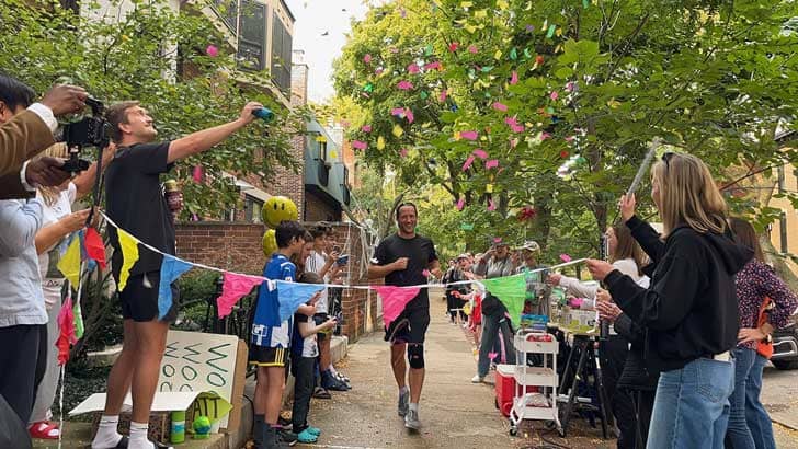 A person wearing a black shirt, shirts, and running shoes running up to a "finish line" with flags on it, people stand on either side of the finish line cheering and throwing confetti