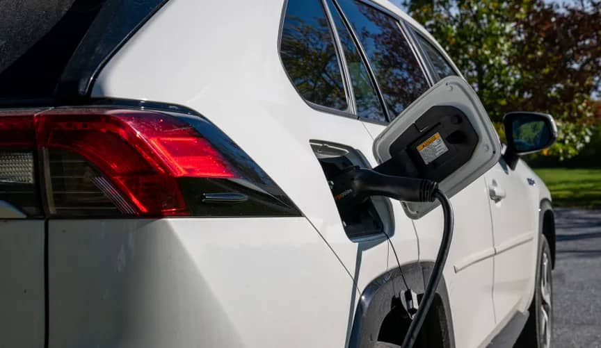 White SUV plugged into a charging station with the charging cable connected to the vehicle's port. Red taillights and green foliage visible in the background.