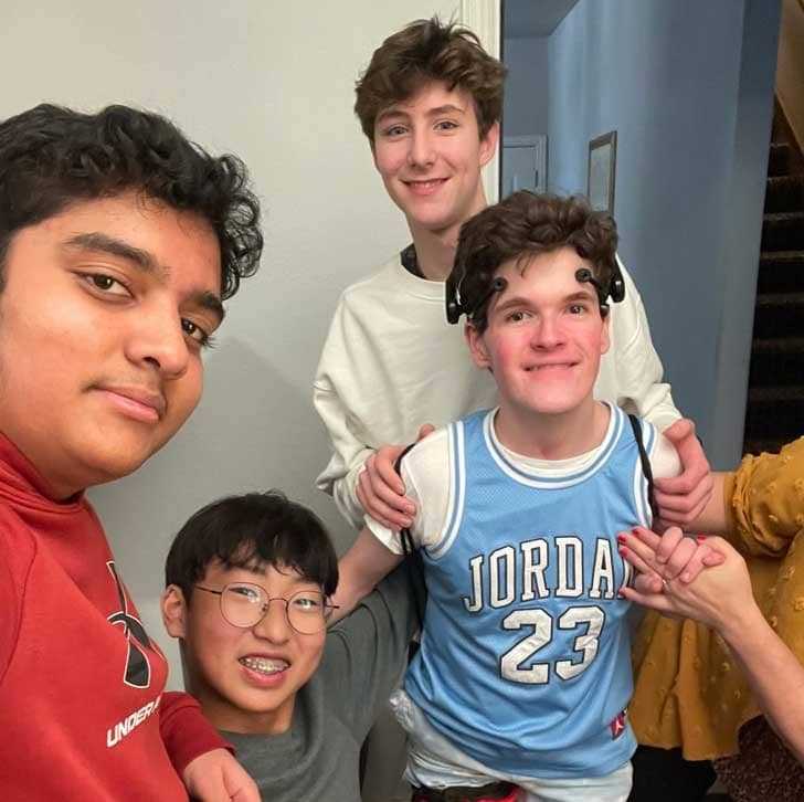 Four teenage boys, three standing around their friend wearing their device