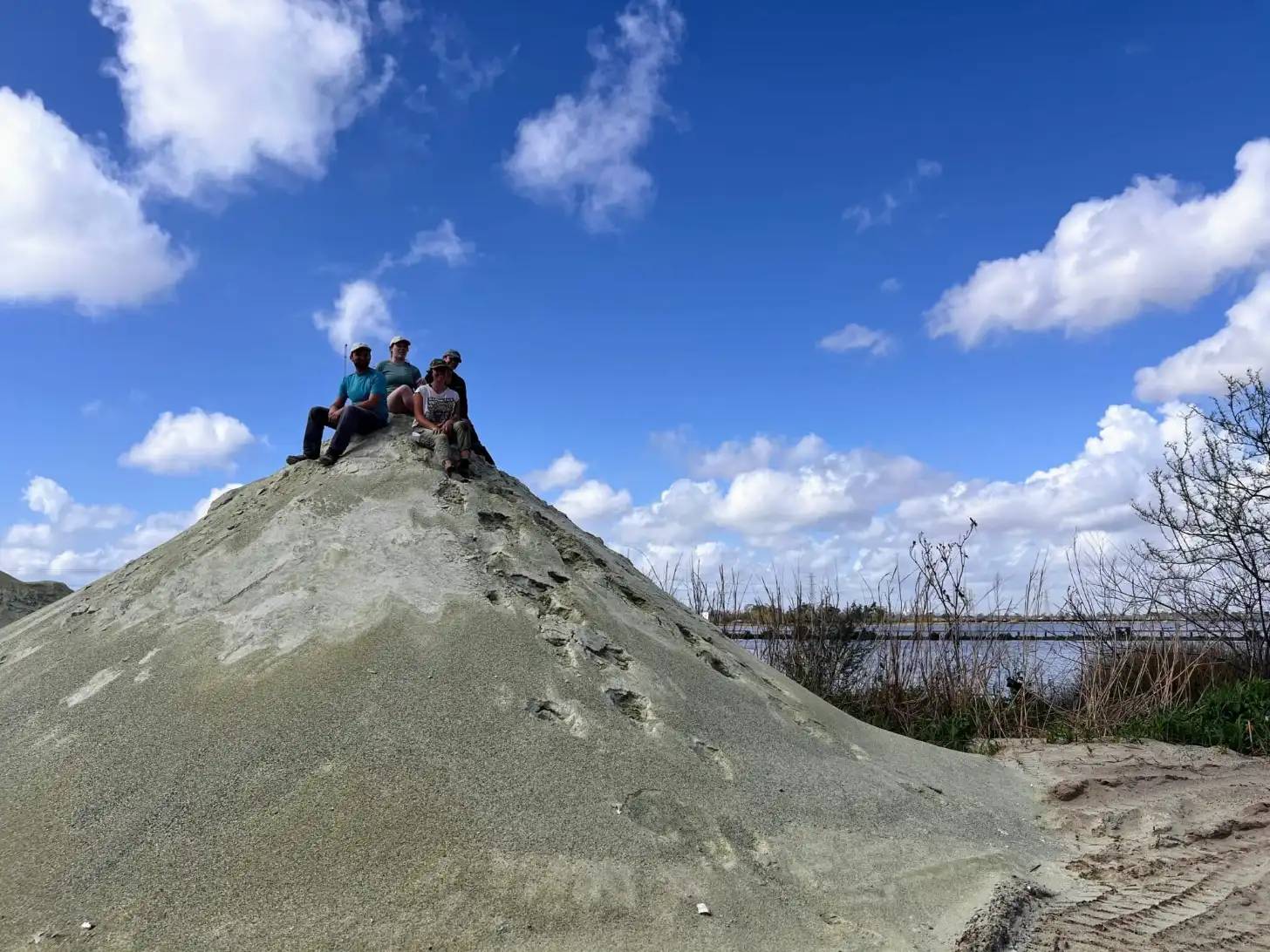 Four people sit atop a large conical mound of light gray sand under a blue sky with white clouds. A body of water and bare vegetation are visible in the background.