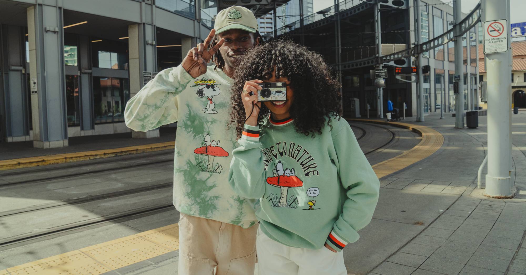 Two people standing next to train tracks in an urban environment wearing green-colored Peanuts Parks Project merchandise