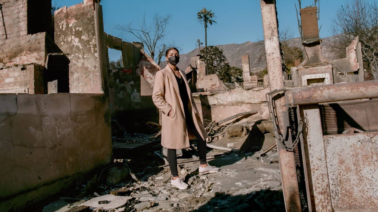 A person wearing a beige coat and face mask stands among ruins of concrete and brick buildings, with mountains visible in the background under clear sky.