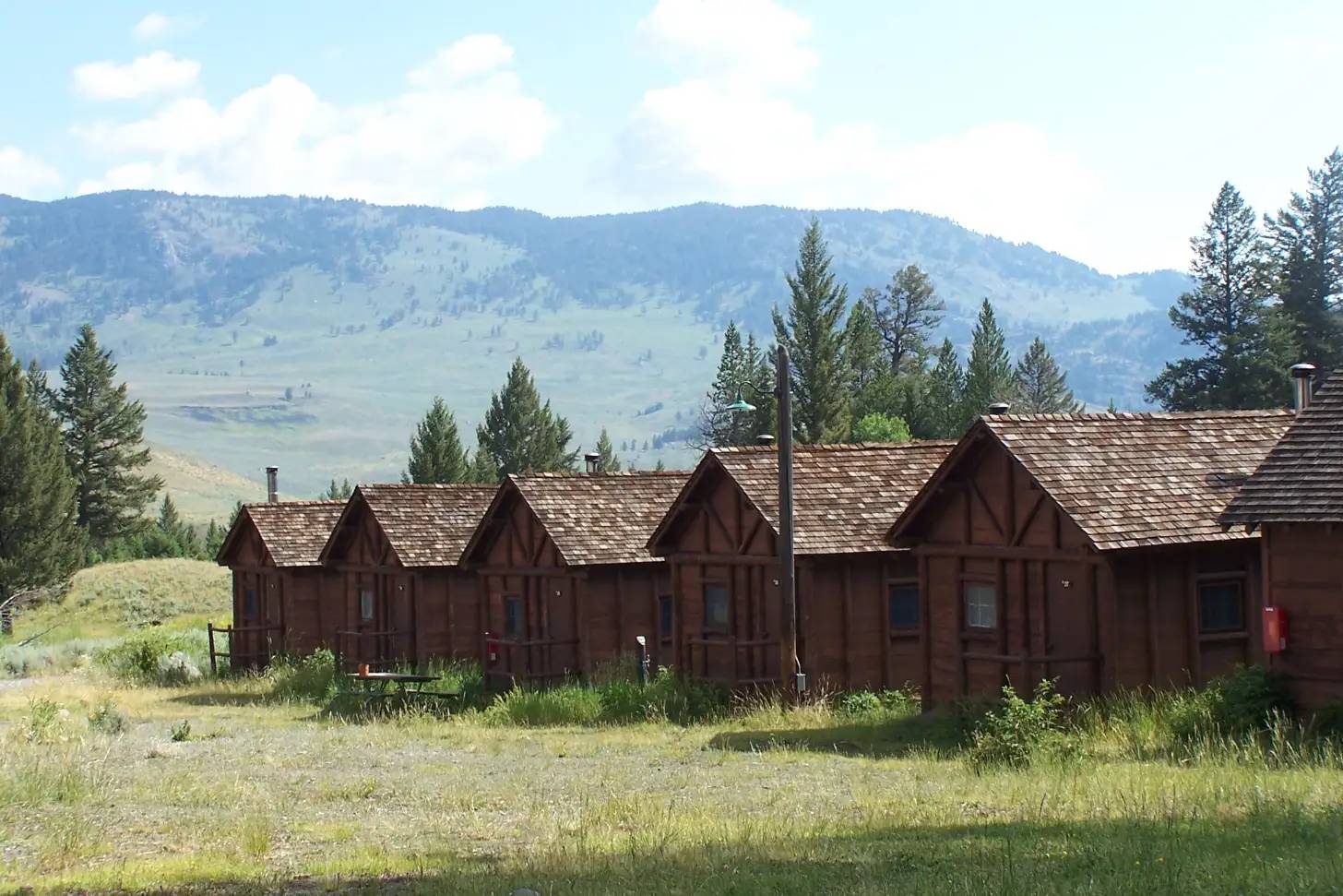 Row of dark brown wooden cabins with peaked roofs set against forested mountains and rolling hills under partly cloudy sky.
