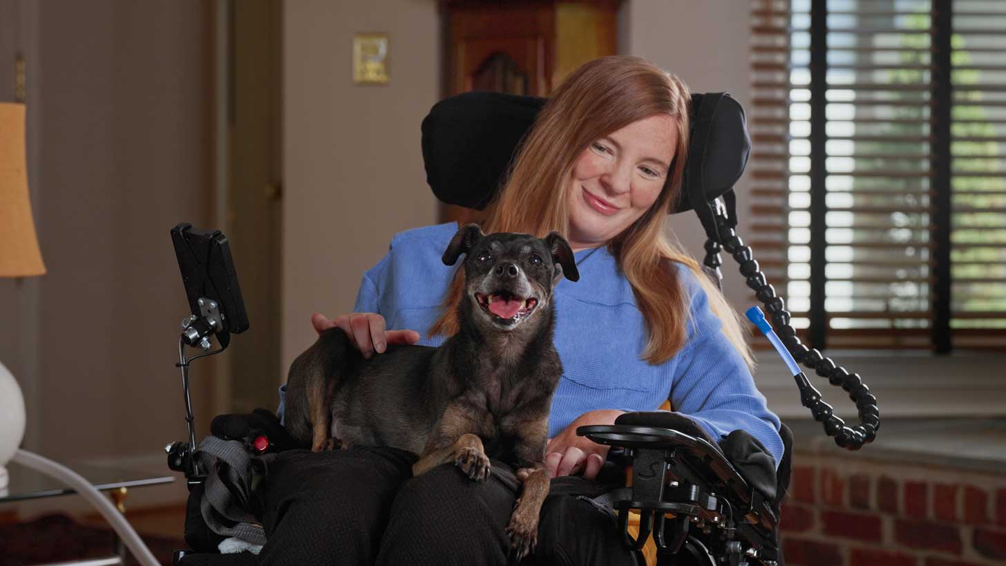 A black and brown small dog sitting on the lap of a woman in a wheelchair, she is petting him