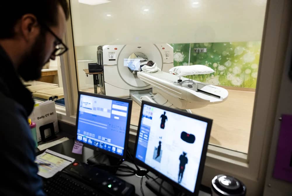 A doctor looking at two screen behind a window where a person is getting a scan in the other room
