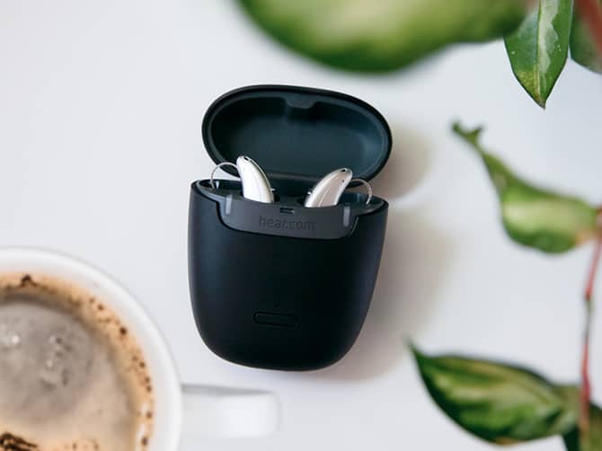 A pair of silver hearing aids inside a black flip-top case with a plant and cup of coffee next to it