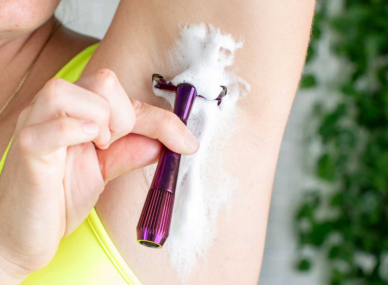 Close-up of a person's armpit with white foamy shaving cream and a hand holding a purple metal Leaf razor 