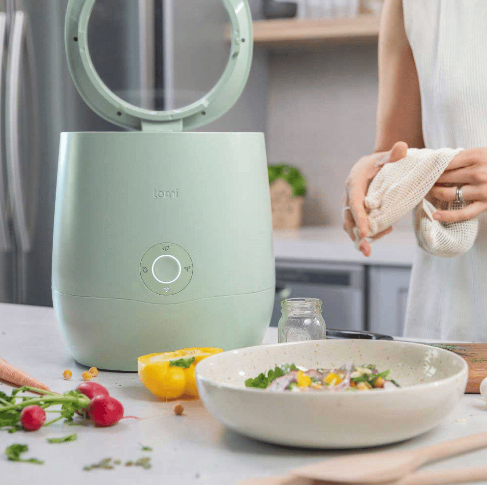 Light green composter on a countertop next to a person making a dish with vegetables
