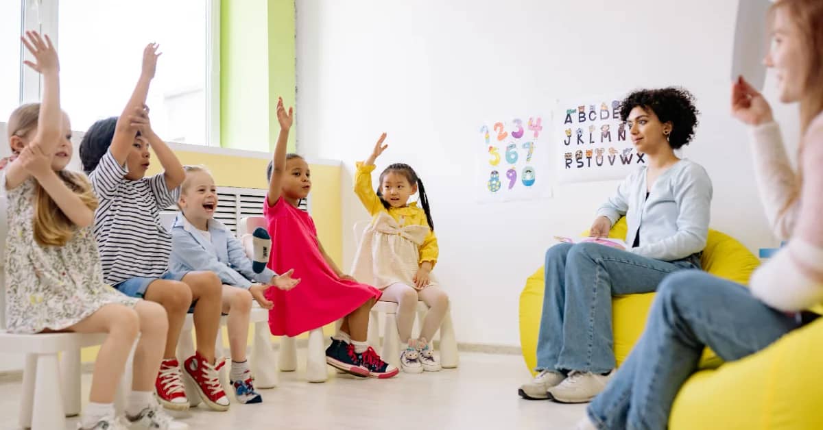 Five children on stools in a classroom setting, two teachers are on yellow bean bags in front of them