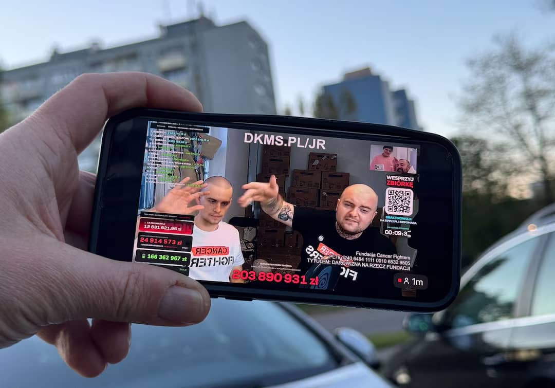 A person holding their phone with two people with shaved heads pictured during a livestream on the screen