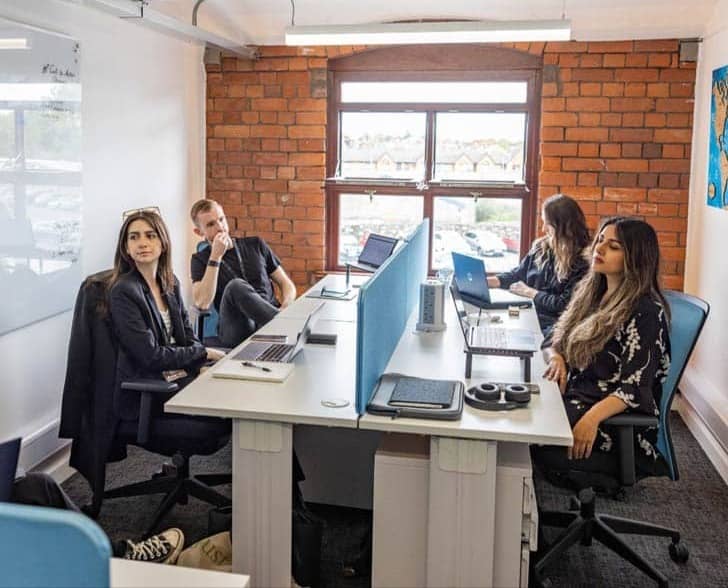 Four people sitting around a shared desk space in a room with a brick wall and window