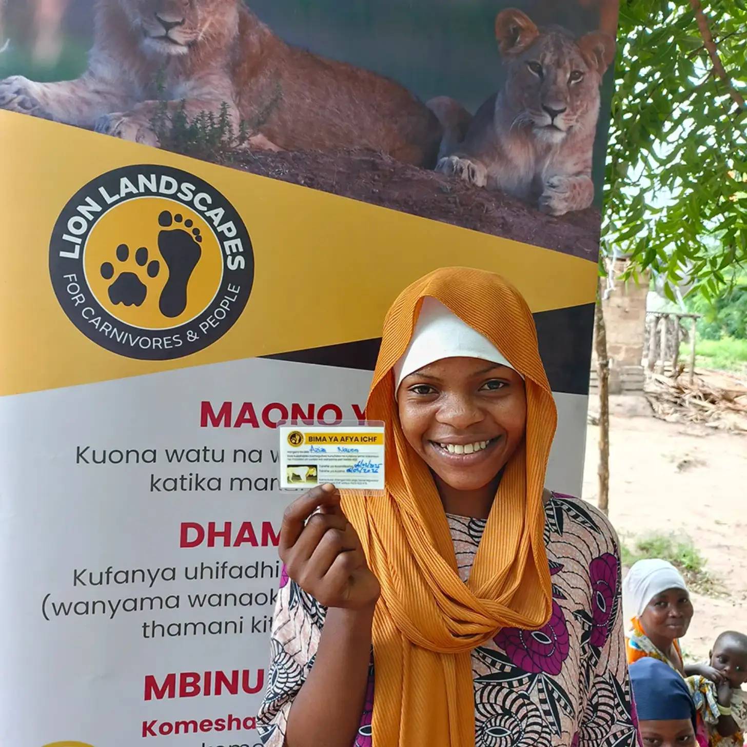 A smiling girl wearing an orange headscarf holds up an identification card in front of a yellow and black banner that reads "LION LANDSCAPES FOR CARNIVORES & PEOPLE" with a paw print logo. Large lions are visible in an image at the top of the banner.