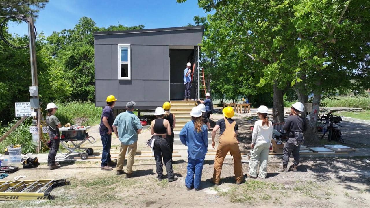 Students standing around the construction site of a black-exterior tiny home