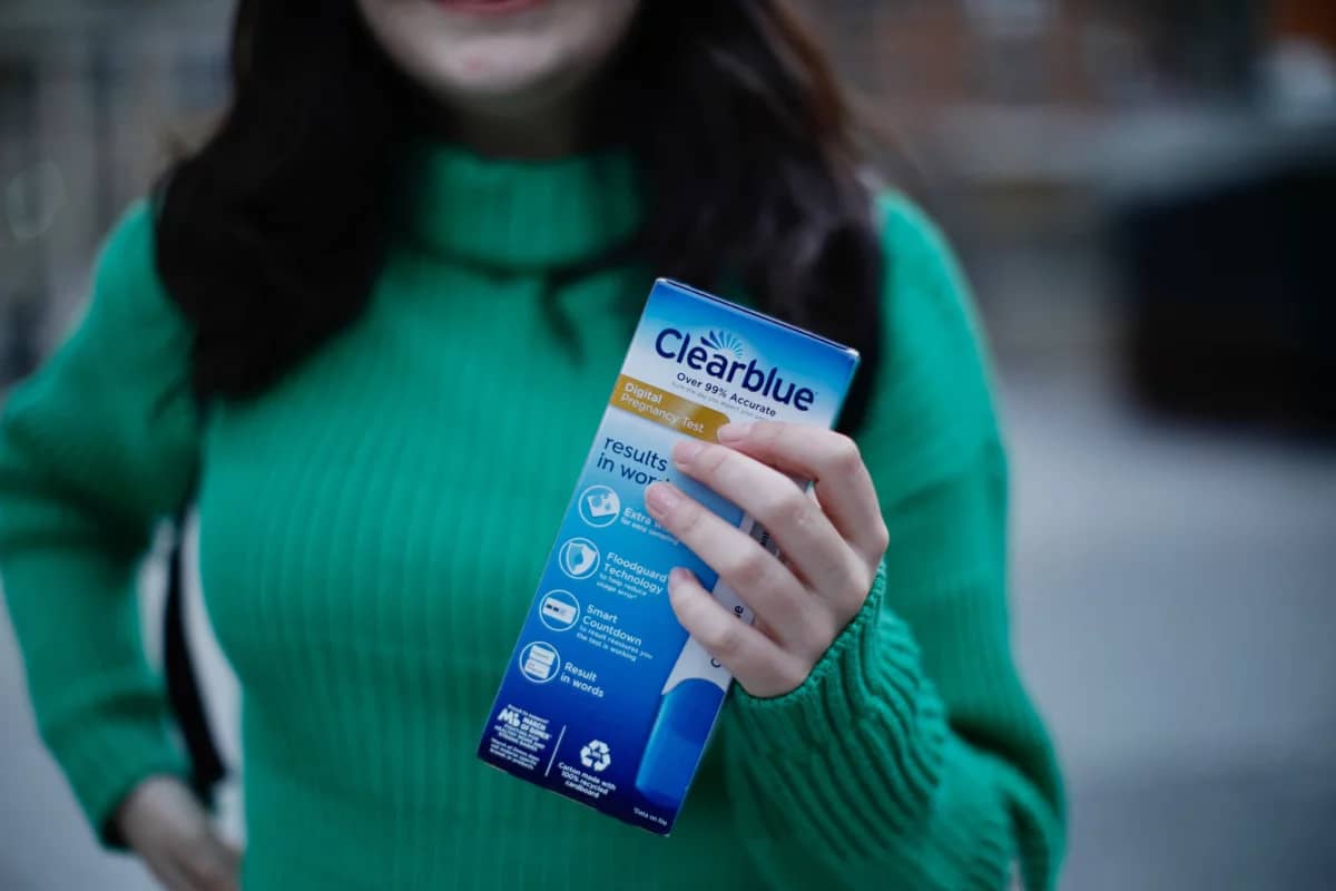 A person wearing a green sweater holding a blue pregnancy test box