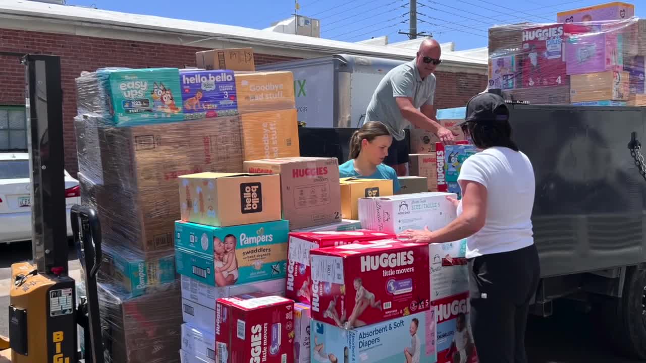 Three people load boxes of diapers and baby products including Huggies and Pampers brands onto a trailer outside a brick building on a sunny day.