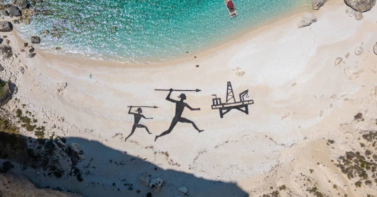 Aerial photo of a beach with black figures of two people holding spears aimed at an oil rig, the artwork is just offshore