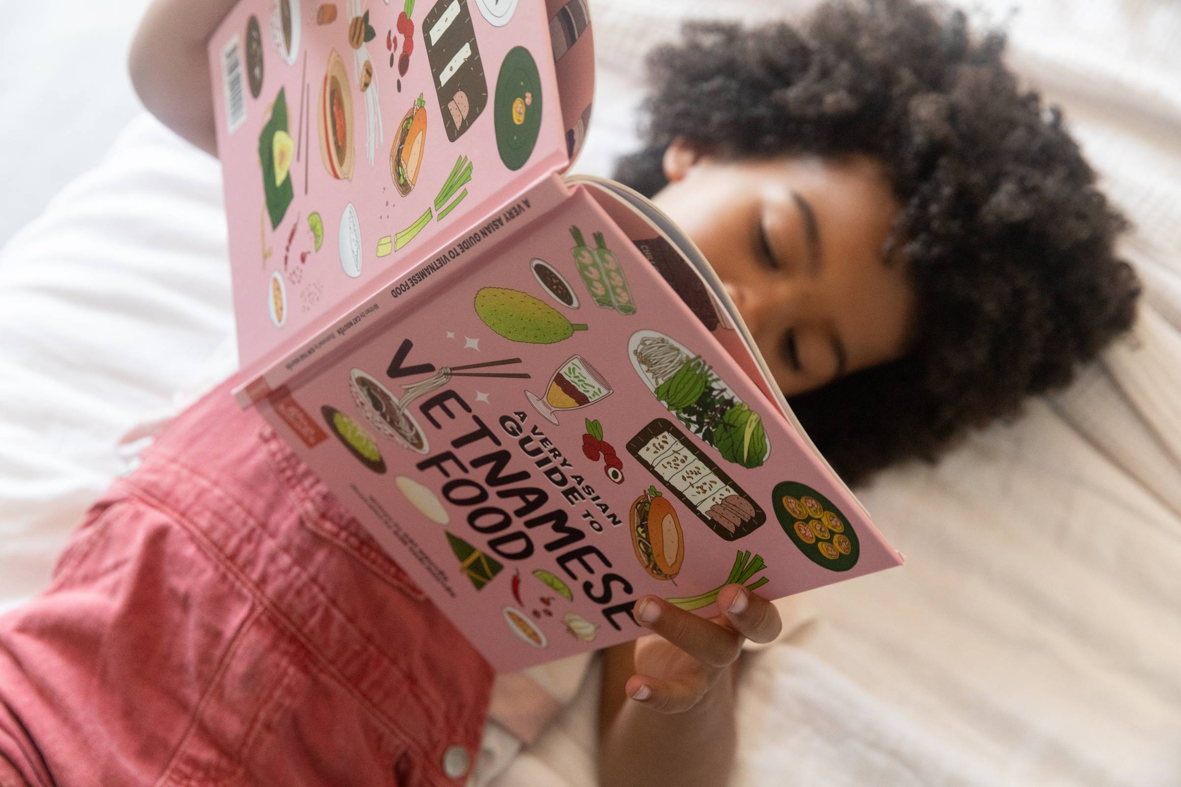 A young girl wearing red overalls laying on her bed reading a book titled "A Very Asian Guide to Vietnamese Food"