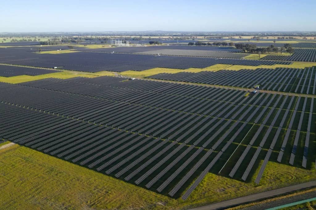 Aerial view of large solar panel arrays arranged in rows across flat agricultural land, with green fields and industrial buildings visible in the distance under clear sky.