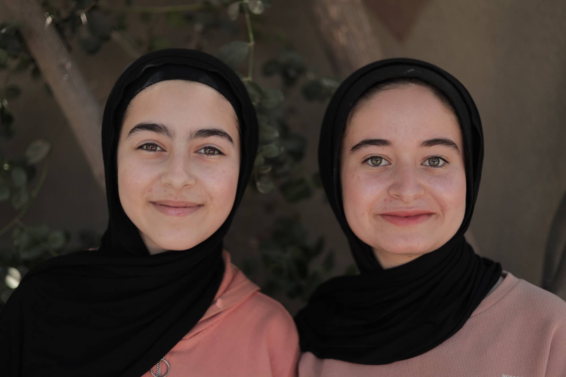 Two young women wearing black headscarves and colored long-sleeved shirts smile at the camera against a neutral background with ivy visible behind them.