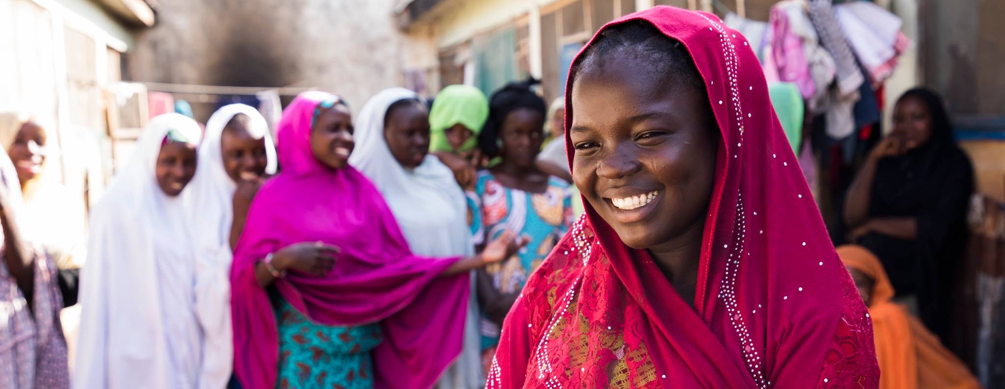 A girl wearing a magenta pink headscarf, smiling, with a larger group of people in the background